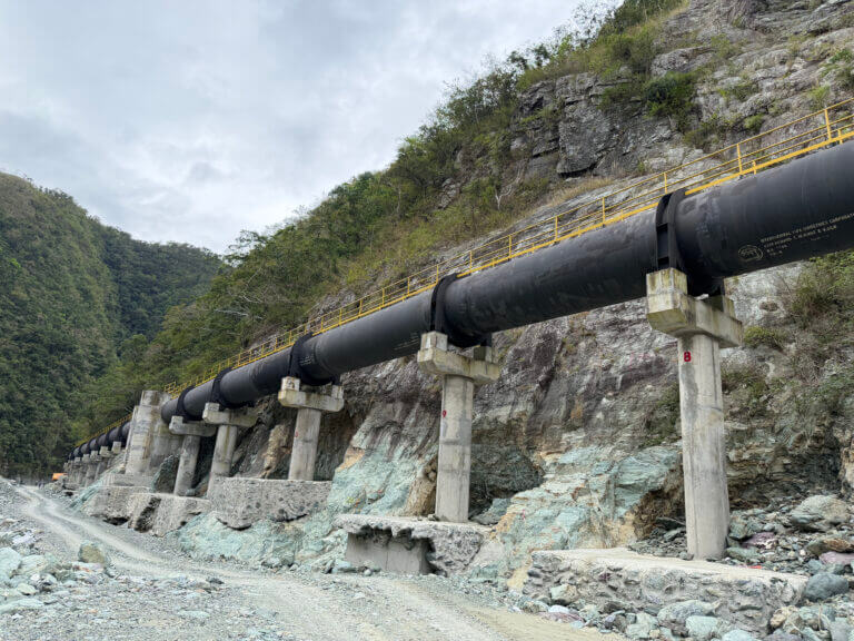 Long black pipeline mounted on concrete supports running along a rocky hillside with mountains and cloudy sky in the background.