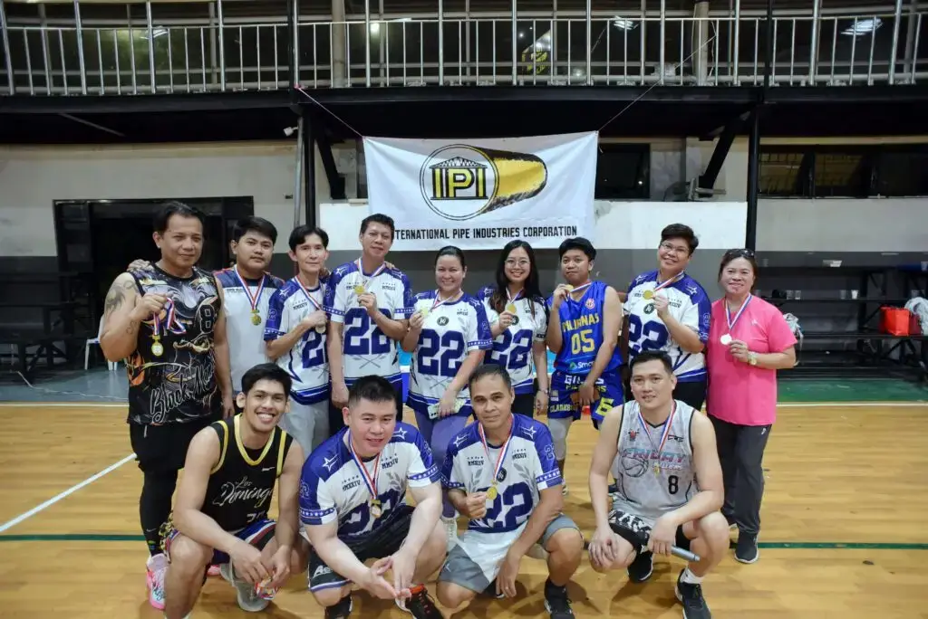 Group of amateur basketball players posing on an indoor court: a mixed group of about 14 people — men and women — wearing matching white-and-blue jerseys and shorts (some kneeling in front, others standing), smiling toward the camera with a tournament banner hanging on the railing behind them.
