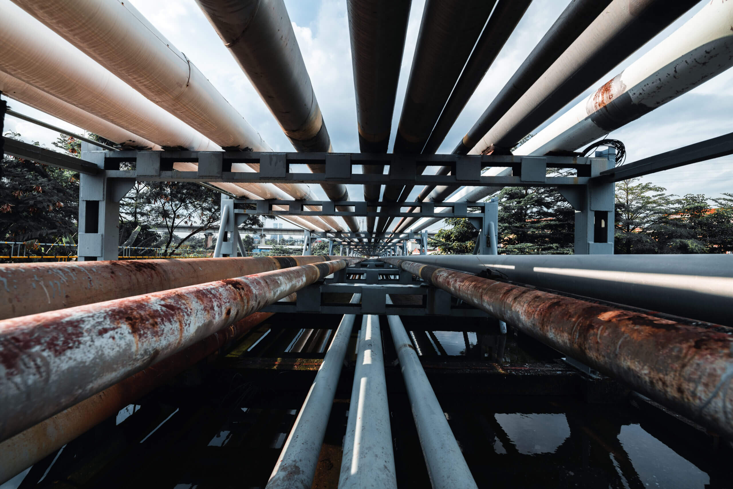 Low-angle view down the center of an industrial structure: symmetrical steel beams and pipes converge toward a vanishing point over a narrow water channel, with reflections of the metal and hints of rust and sky visible.