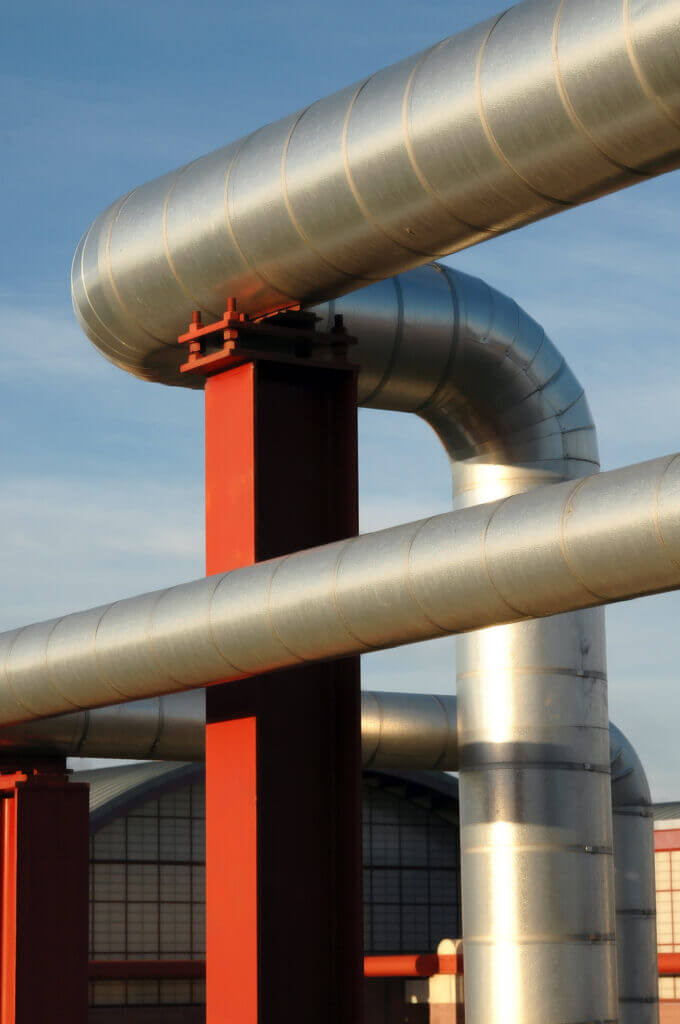 Close-up of large insulated industrial pipes: shiny curved metal ducts cross in front of a red vertical support column against a pale blue sky.