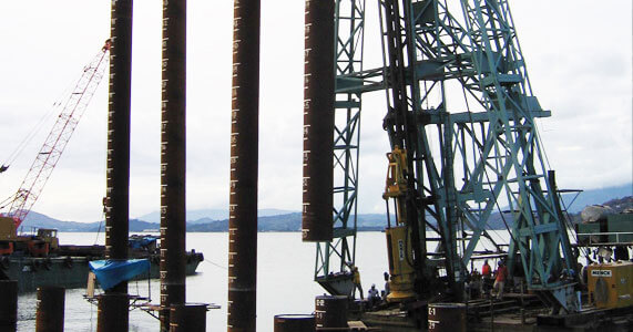 Close-up view of a marine construction or bridge-support structure: tall vertical steel piles driven into the water with rigging and scaffolding, and a worker operating equipment on a platform.