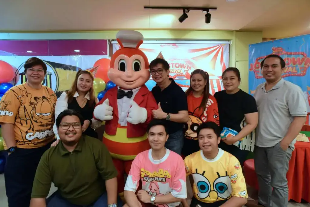 Group photo of eight people posing indoors around a large costumed character (bright cartoon mascot wearing red and yellow); some people are seated in front while others stand behind the mascot, and a decorated backdrop with party signage is visible.
