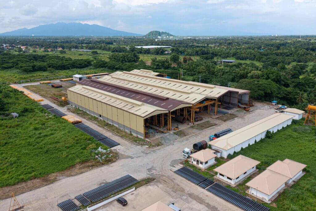 Aerial view of an industrial complex: multiple long factory buildings with rust-colored roofs, surrounding roads, parking areas and green fields extending to low hills on the horizon.
