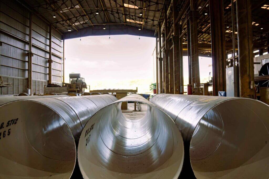 Large industrial warehouse interior with several massive shiny metal pipes or tubes lying on the floor, seen from pipe-end level toward the open doorway and bright daylight outside.