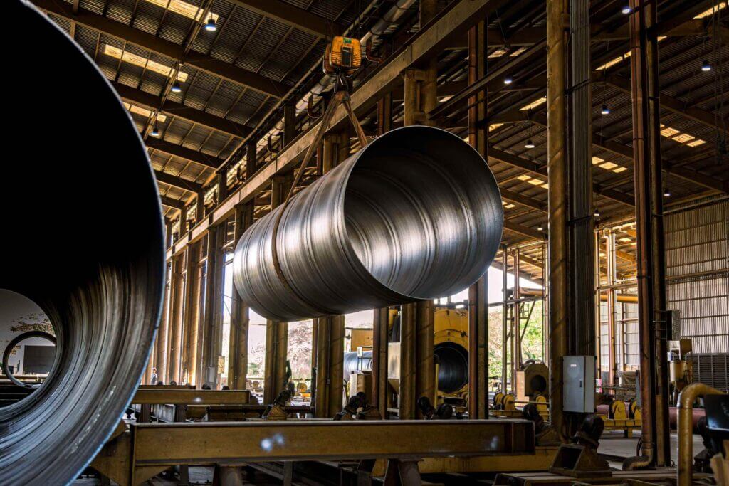 Close-up inside a factory showing a large cylindrical metal pipe suspended by a crane hook inside a high-roofed production hall, with rows of vertical supports and equipment in the background.