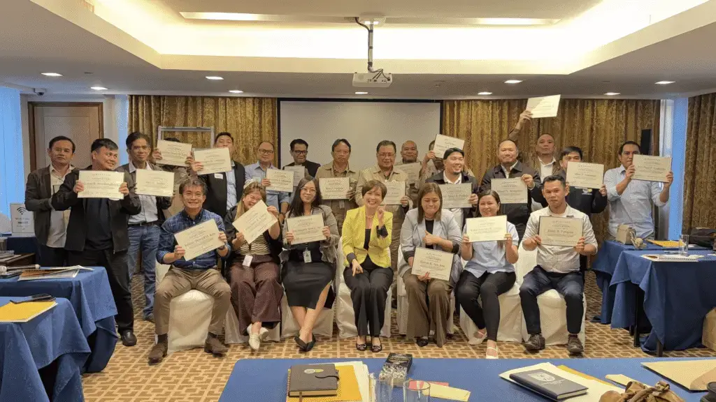 About twenty people arranged in three rows in a conference room, many holding certificates toward the camera; patterned curtains and a projection screen are visible behind them, and a table with papers sits in the foreground.