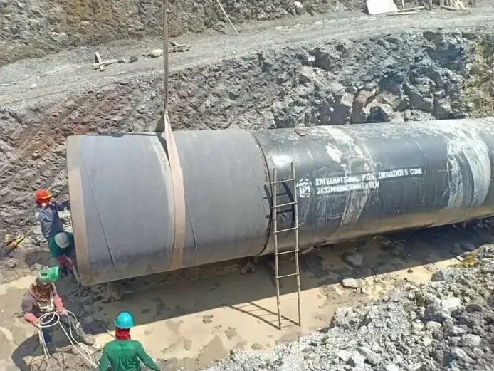 Close-up of a large welded steel pipe segment resting on gravel with a metal ladder leaning against it and a worker wearing a green hard hat nearby; rocky slope in the background.