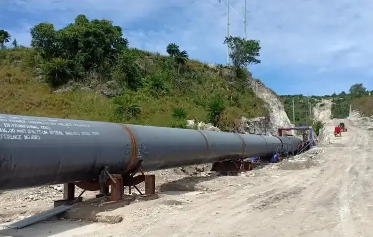 Long steel pipeline sections laid on supports along a dirt road at the base of a green hillside; workers and equipment visible in the distance under a partly cloudy sky.